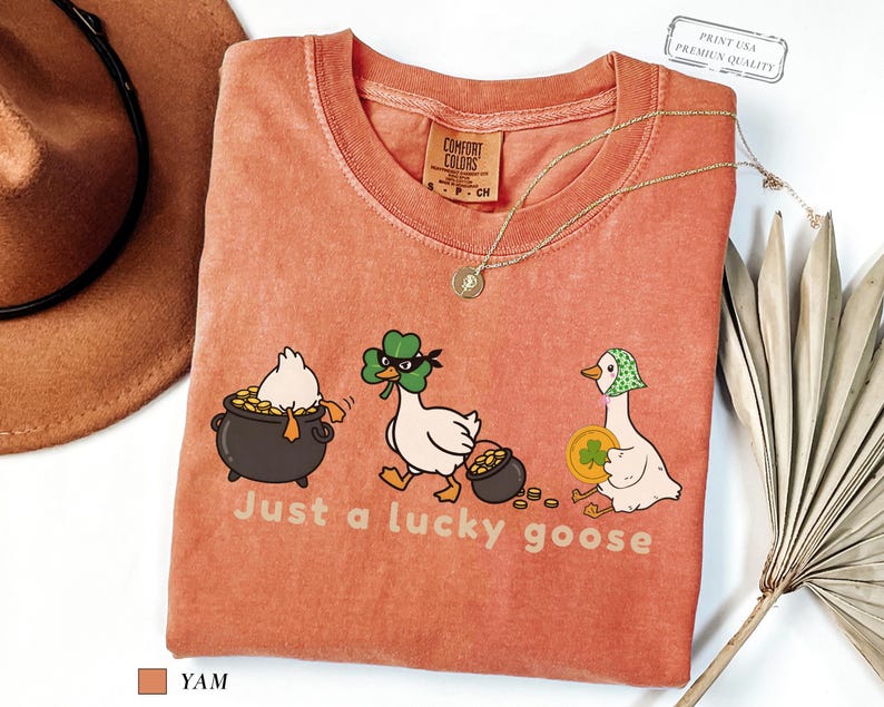 yam comfort colors t-shirt with three illustrated geese, one in a pot of gold, one wearing a shamrock mask, and one holding a coin, playful st patrick’s day design