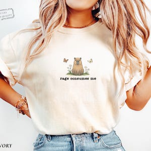 Ivory Comfort Colors® t-shirt featuring a vintage capybara illustration sitting in grass with small flowers and butterflies, with the phrase “rage consumes me”.