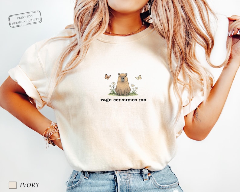 Ivory Comfort Colors® t-shirt featuring a vintage capybara illustration sitting in grass with small flowers and butterflies, with the phrase “rage consumes me”.