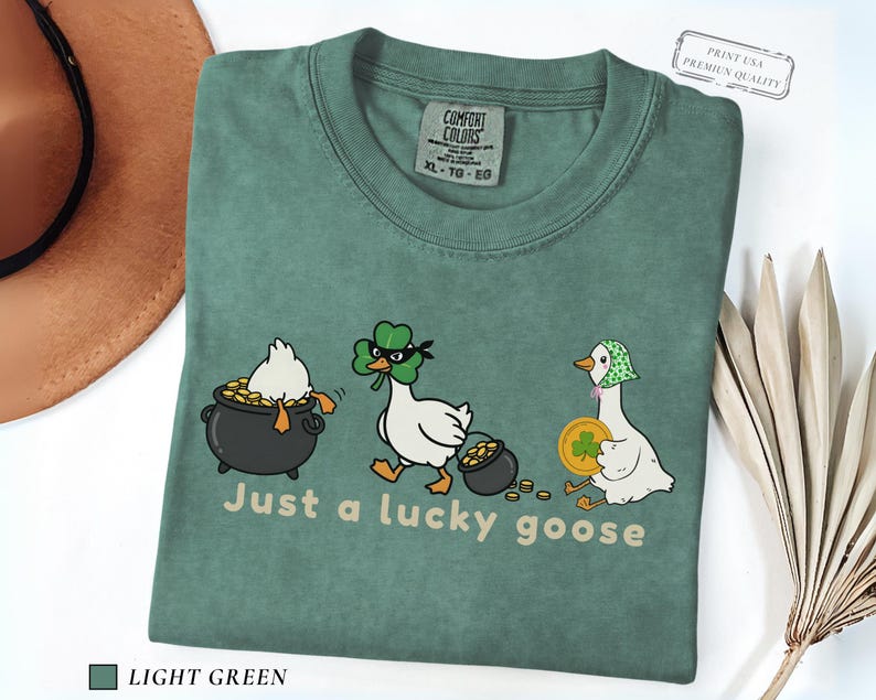 light green comfort colors t-shirt with three illustrated geese, one in a pot of gold, one wearing a shamrock mask, and one holding a coin, playful st patrick’s day design