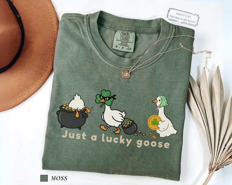 moss comfort colors t-shirt with three illustrated geese, one in a pot of gold, one wearing a shamrock mask, and one holding a coin, playful st patrick’s day design