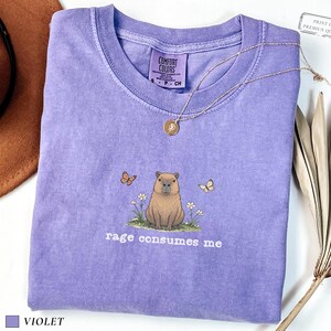 Violet Comfort Colors® t-shirt featuring a vintage capybara illustration sitting in grass with small flowers and butterflies, with the phrase “rage consumes me”.