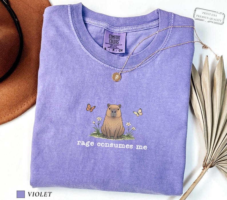 Violet Comfort Colors® t-shirt featuring a vintage capybara illustration sitting in grass with small flowers and butterflies, with the phrase “rage consumes me”.