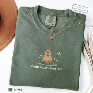 Moss Comfort Colors® t-shirt featuring a vintage capybara illustration sitting in grass with small flowers and butterflies, with the phrase “rage consumes me”.