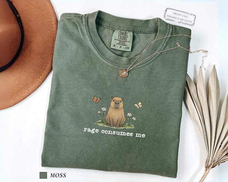 Moss Comfort Colors® t-shirt featuring a vintage capybara illustration sitting in grass with small flowers and butterflies, with the phrase “rage consumes me”.