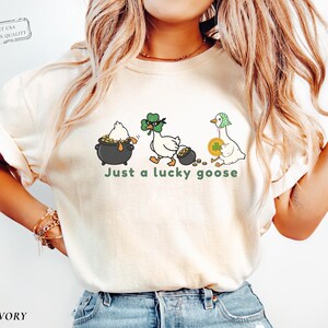 ivory comfort colors t-shirt with three illustrated geese, one in a pot of gold, one wearing a shamrock mask, and one holding a coin, playful st patrick’s day design