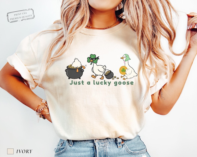 ivory comfort colors t-shirt with three illustrated geese, one in a pot of gold, one wearing a shamrock mask, and one holding a coin, playful st patrick’s day design