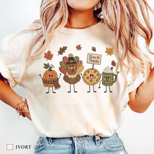 May include: An ivory short-sleeve t-shirt featuring a graphic print of a cartoon pumpkin, a turkey wearing a hat, a pie with a sign that reads Give Thanks, and a mug with whipped cream, all surrounded by various autumn leaves and acorns.