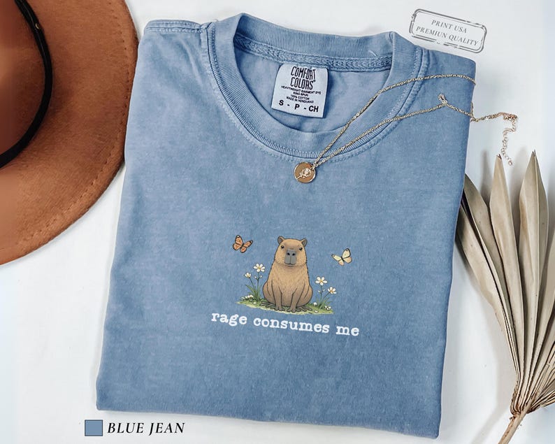 Blue Jean Comfort Colors® t-shirt featuring a vintage capybara illustration sitting in grass with small flowers and butterflies, with the phrase “rage consumes me”.
