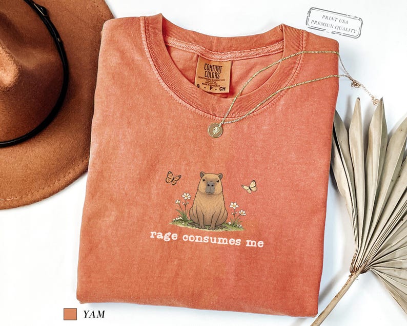 Yam Comfort Colors® t-shirt featuring a vintage capybara illustration sitting in grass with small flowers and butterflies, with the phrase “rage consumes me”.