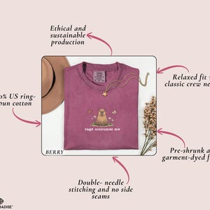 May include: Berry-colored t-shirt with a crew neckline. The shirt features an embroidered design of a small animal with the text "rage consumes me." The shirt is made of 100% US ring-spun cotton and has double-needle stitching.