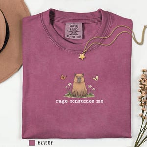 Berry Comfort Colors® t-shirt featuring a vintage capybara illustration sitting in grass with small flowers and butterflies, with the phrase “rage consumes me”.
