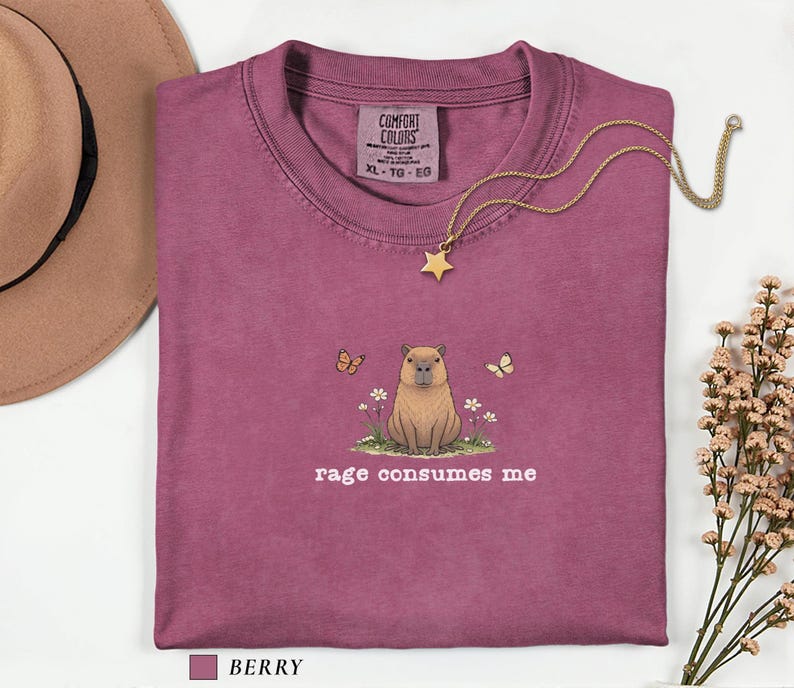 Berry Comfort Colors® t-shirt featuring a vintage capybara illustration sitting in grass with small flowers and butterflies, with the phrase “rage consumes me”.