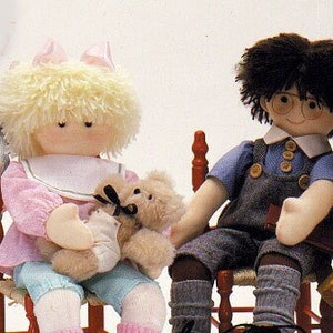 May include: Three handmade rag dolls sit on small wooden chairs. One doll wears a pink outfit and holds a teddy bear. Another doll wears overalls and glasses. The third doll wears a yellow dress and reads a book.