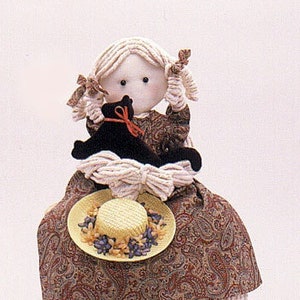 Puede incluir: Una muñeca de trapo con el pelo rubio y un sombrero floral se sienta en un taburete de madera con flecos blancos. La muñeca sostiene un gato negro.