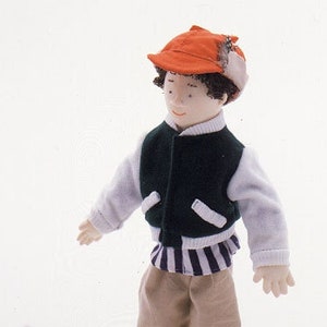 May include: A doll wearing a green and white striped jacket, a red and white baseball cap, and khaki pants. The doll is holding a white stuffed dog with a red and white plaid bow.