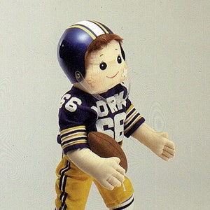 May include: A plush doll wearing a purple and yellow football uniform with a blue helmet and holding a brown football. The uniform has the word "YORK" on the front.