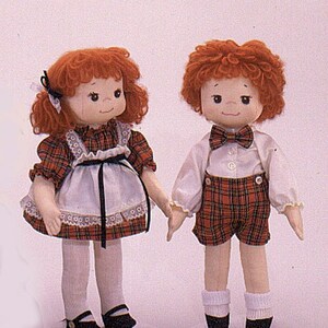 May include: Two red-haired dolls wearing plaid outfits. The girl doll is wearing a white dress with a black sash and lace trim. The boy doll is wearing a white shirt with a plaid waistcoat and shorts. Both dolls have red hair and are smiling.