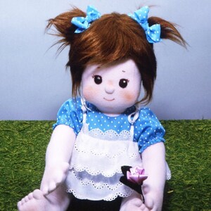May include: A handmade doll with brown hair, blue eyes, and a white dress with blue polka dots. The doll is wearing blue bows in her hair and holding a pink flower.