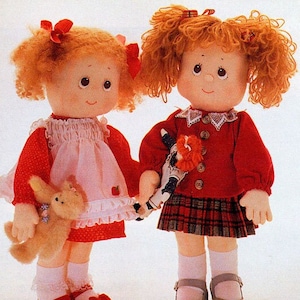 May include: Two red-haired dolls with rosy cheeks and big eyes are holding hands. One doll is wearing a red dress with white polka dots and a pink apron. She is holding a brown plush bunny. The other doll is wearing a red sweater with a white collar and a plaid skirt. She is holding a smaller doll.