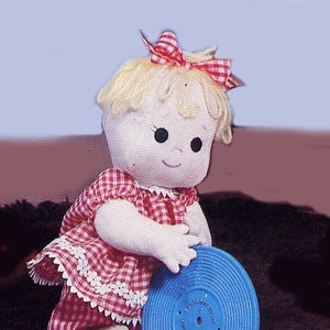 Pode incluir: Uma boneca feita à mão com cabelo loiro, vestindo um vestido xadrez vermelho e branco e um laço vermelho. A boneca está segurando um toca-discos de plástico azul.
