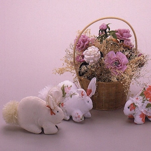 以下が含まれることがあります： 白い毛足の長いウサギのぬいぐるみが3匹、花柄の布で飾られています。1匹はふわふわのしっぽが付いています。薄いグレーの表面に置かれています。ウサギの後ろには、ピンクと白の花が詰まった籐のバスケットがあります。