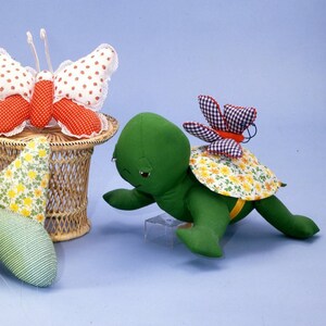 Pode incluir: Três brinquedos de pelúcia feitos à mão: uma tartaruga verde com uma concha amarela e uma pequena borboleta nas costas, uma borboleta verde e amarela com flores brancas e uma borboleta vermelha e branca com bolinhas brancas.
