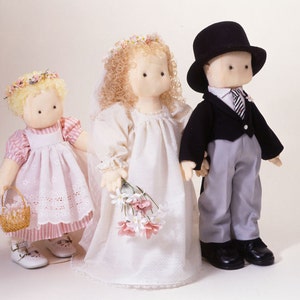 May include: Three fabric dolls dressed in wedding attire. The bride doll is wearing a white dress with a floral bouquet, the groom doll is wearing a black suit with a top hat, and the flower girl doll is wearing a pink and white dress with a basket of flowers.