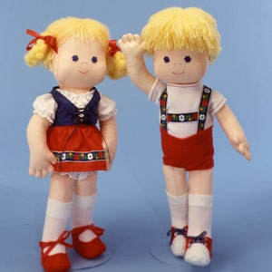 May include: Two handmade dolls dressed in traditional German clothing. The girl doll wears a red and blue dress with a white apron and white socks and red shoes. The boy doll wears red shorts with suspenders and a white shirt with a red and white striped band. Both dolls have blonde hair and blue eyes.