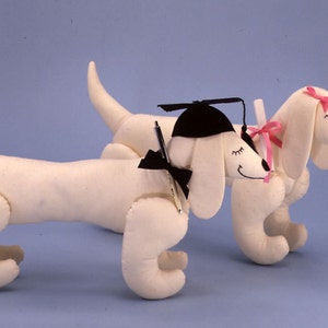 Pode incluir: Dois cães salsicha de pelúcia brancos usando toucas de formatura e um laço rosa. O cão à esquerda está segurando uma caneta preta e tem uma gravata borboleta preta. O cão à direita tem um laço rosa na cabeça.