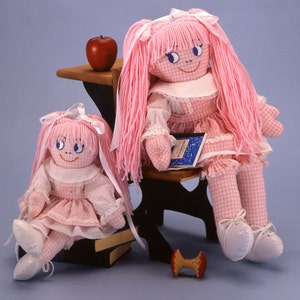 May include: Two pink fabric dolls with long pink hair and pink and white gingham dresses. The larger doll is sitting on a wooden chair with a book in her lap. The smaller doll is sitting on a stack of books. Both dolls have blue eyes and smiles.