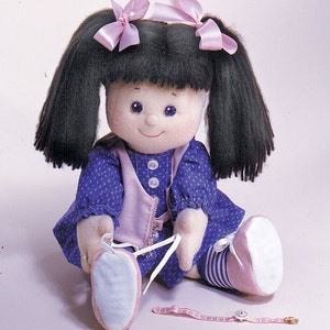 May include: A handmade doll with black hair in pigtails, wearing a blue and white patterned dress with a pink vest and white shoes. The doll is holding a pink and white ribbon in its hands.