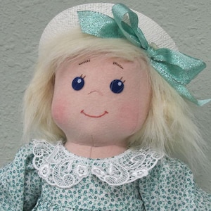 May include: A handmade doll with blonde hair, wearing a white hat with a green bow, a white lace collar, and a green floral dress.