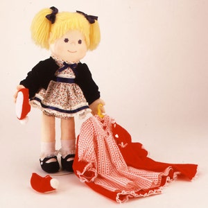 Peut inclure: Une poupée aux cheveux jaunes portant un cardigan noir, une robe à fleurs et des chaussures noires. La poupée tient une chaussure rouge dans une main et une robe rouge sur un cintre dans l'autre.