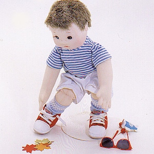 May include: A cloth doll with brown hair wearing a blue and white striped shirt, white shorts, red shoes, and blue and white socks. The doll is holding a pair of red sunglasses and a blue toy.