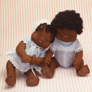 May include: Two handmade cloth dolls with brown skin, black hair, and blue and white polka dot dresses. The dolls are sitting on a pink and white striped background.