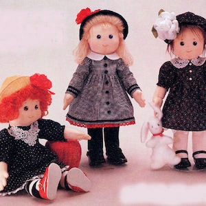 May include: Three handmade fabric dolls. One doll is seated, wearing a black dress with white polka dots, a yellow hat with red trim, and red shoes. Another doll stands in a black and white checkered dress. The third doll holds a white stuffed bunny.