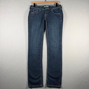 May include: A pair of dark blue denim jeans featuring a classic five-pocket design, a button and zipper fly closure, belt loops, and subtle whiskering details on the front thighs. The jeans have a straight-leg cut and a mid-rise waistline.