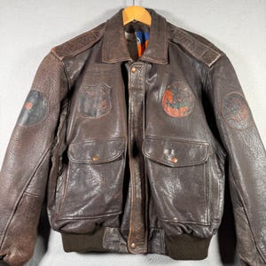 Puede incluir: Una chaqueta bomber de cuero marrón oscuro con cuello clásico, dos bolsillos delanteros con solapa y cierre de broche, y un dobladillo de punto acanalado. La chaqueta está adornada con varios parches circulares y en forma de escudo.