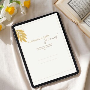 Könnte beinhalten: Ein digitales Tablet mit dem Text "TAWAKKUL & SABR Journal" und einem goldenen Blattdesign. Das Tablet liegt auf einem weißen Tuch, zusammen mit einem Buch und Narzissen.
