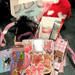 May include: An open gift box filled with various items. Includes a red heart-shaped sock, face masks, a lip gloss, a keychain, a bookmark with a beaver illustration, and stickers with bows, butterflies, and a teddy bear. The box is decorated with pink accents.