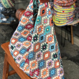 May include: A patterned tote bag with a brown handle. The bag features a geometric design in shades of orange, blue, green, and red on a grey background. The bag is sitting on a wooden bench.