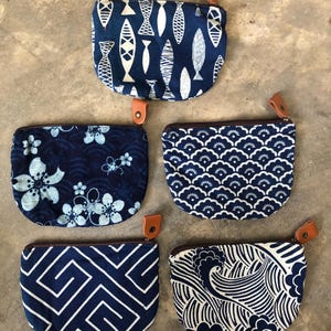 Könnte beinhalten: Fünf halbkreisförmige Stoffbeutel mit verschiedenen blau-weißen Mustern, darunter Fisch-, Blumen-, Wellen- und geometrische Designs. Jeder Beutel ist mit einem dunkelbraunen Reißverschluss und einer kleinen, lederartigen Zuglasche ausgestattet.