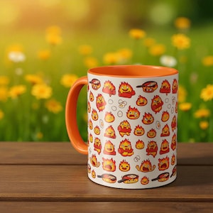 Könnte beinhalten: Eine weiße Keramiktasse mit orangefarbenem Innenraum und Griff, verziert mit Cartoon-Flammen und Kochutensilien. Die Tasse steht auf einer Holzoberfläche mit einem verschwommenen Hintergrund aus gelben Blumen und grünem Laub.