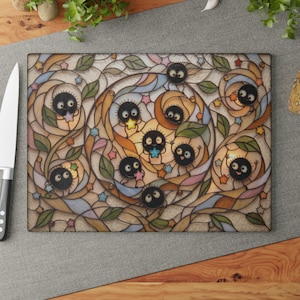 Pode incluir: Tábua de corte retangular de vidro com um design de vitral. Apresenta sprites de fuligem pretos, estrelas e folhas. A tábua tem um fundo bege claro com detalhes em azul, rosa e laranja. Uma faca é visível à esquerda.
