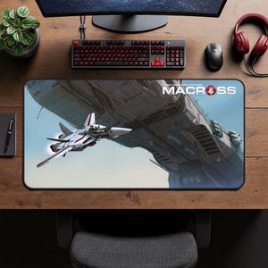 May include: A rectangular mousepad featuring a detailed illustration of a white and red fighter jet in flight, approaching a large spacecraft. The mousepad has the text "SUPER DIMENSION FORTRESS MACROSS" in white.
