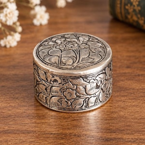May include: A small, round, silver-colored metal box featuring intricate embossed floral and leaf patterns on both the lid and the sides. The container has a polished finish with darkened recessed areas that highlight the detailed botanical engravings.