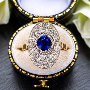 May include: A gold ring with an oval-shaped face, featuring a large, round, deep blue gemstone at the center, surrounded by smaller, clear gemstones. The ring is presented in a black jewelry box with a gold trim.