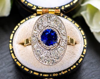 May include: A gold ring with an oval-shaped face, featuring a large, round, deep blue gemstone at the center, surrounded by smaller, clear gemstones. The ring is presented in a black jewelry box with a gold trim.