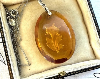 May include: An oval amber pendant necklace with a floral design, displayed in a vintage jewelry box. The pendant is suspended from a silver chain. The box is lined with cream-colored fabric and has a dark brown exterior.
